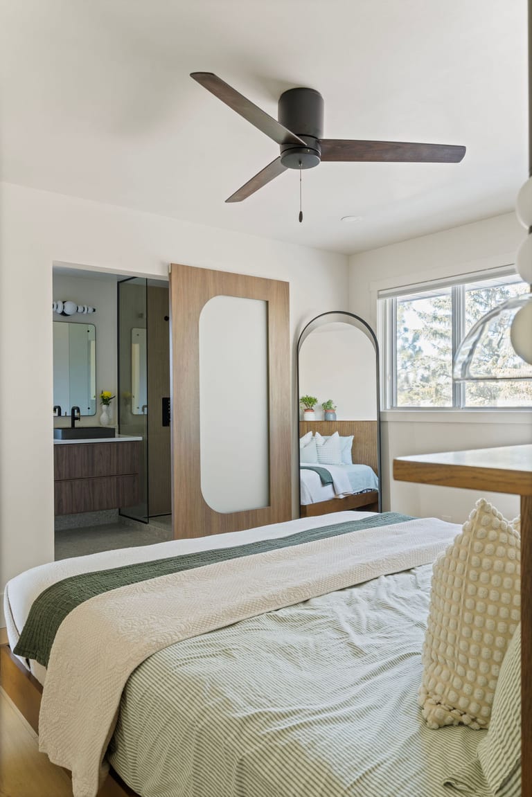 A bright primary bedroom with a ceiling fan, an arched floor mirror and a walnut sliding door open to the ensuite.