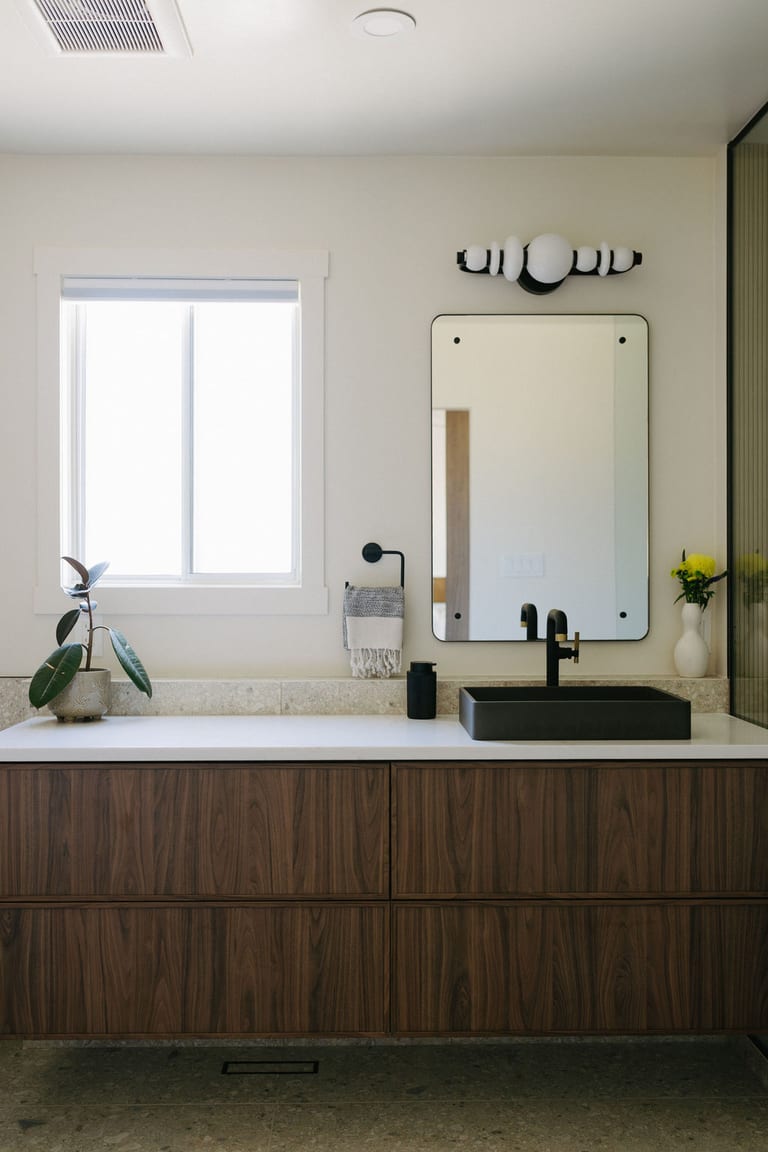 A floating walnut vanity with a white counter, a dark vessel sink, a black faucet and a window.