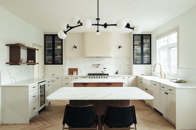 A kitchen with two islands, white stone counters, a full height stone backsplash, a curved range hood and a black globe chandelier.