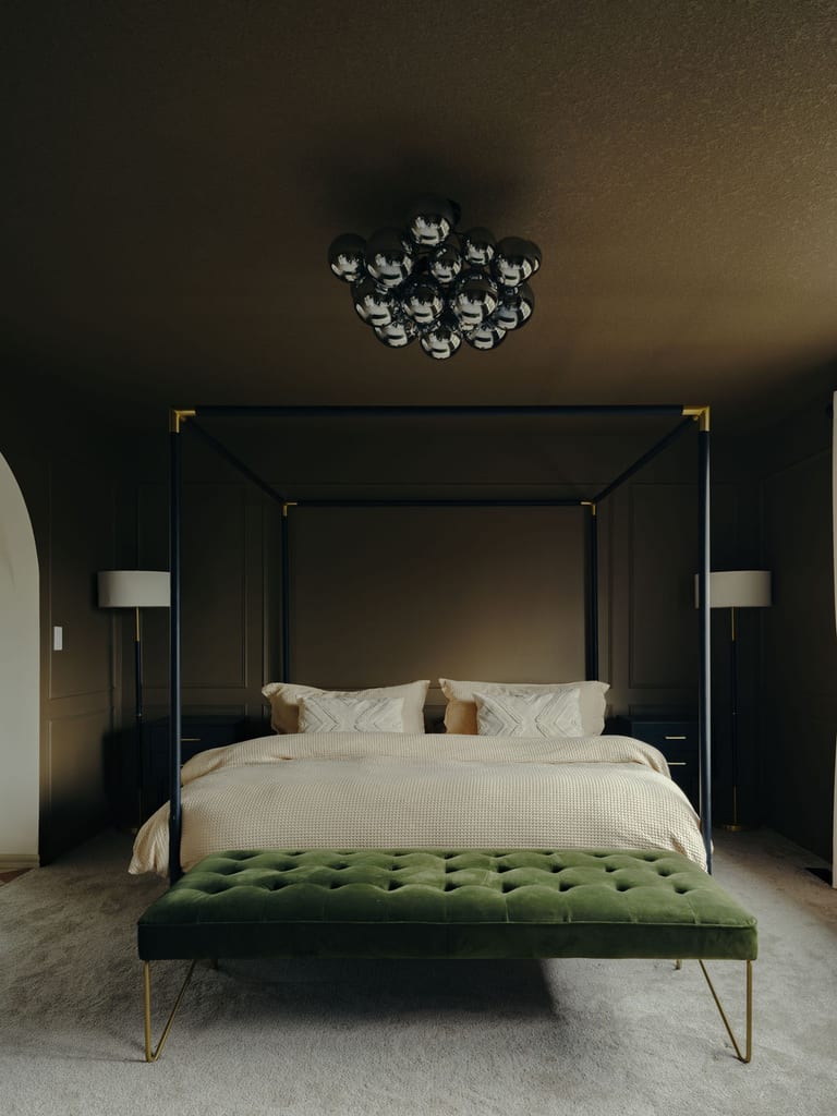 A primary bedroom with dark walls and ceiling, a black canopy bed, a green velvet bench and a modern chandelier.