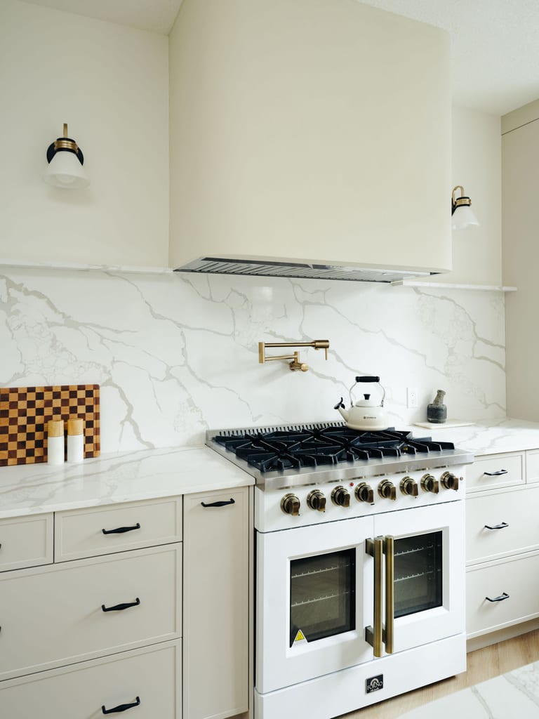 A white range with brass knobs, a brass pot filler and a full height stone backsplash under a curved hood.