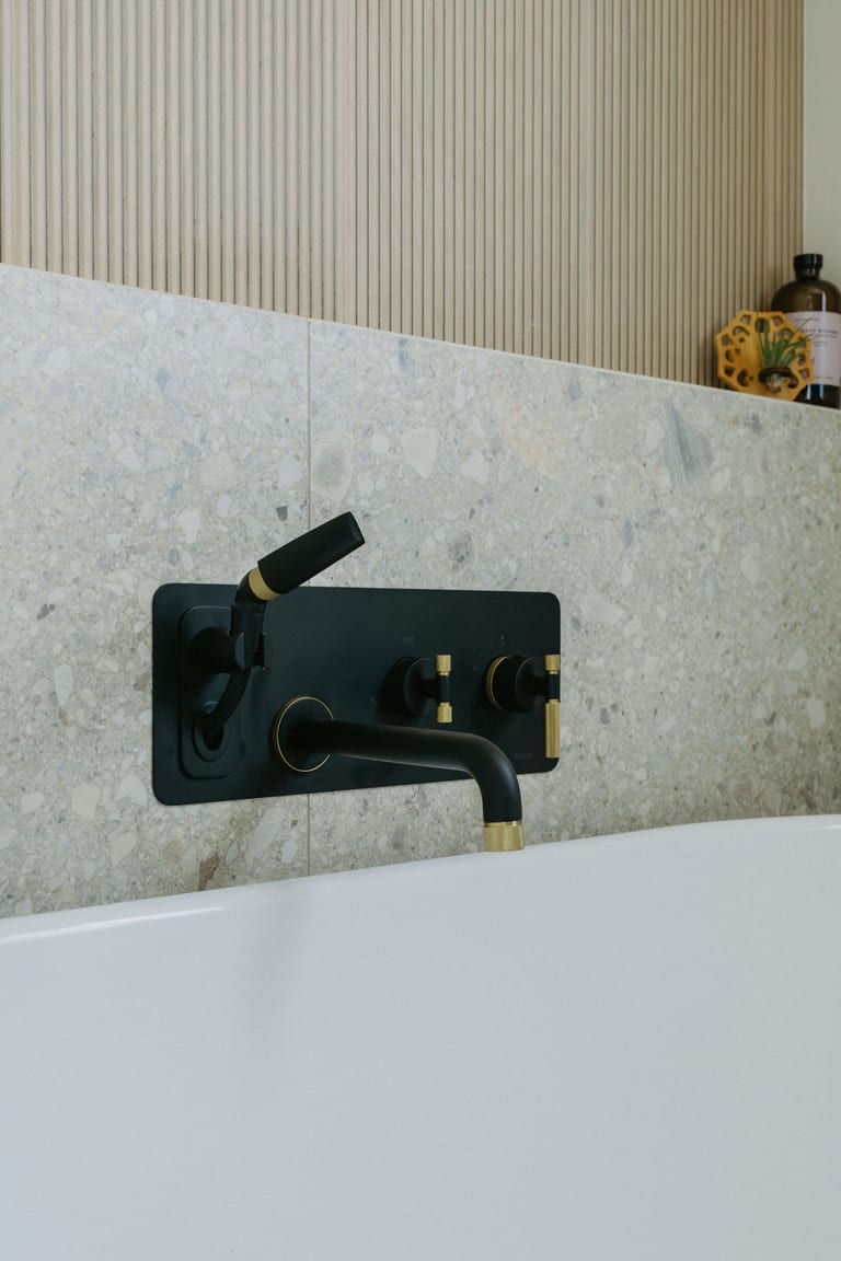 A black and brass wall mounted tub faucet with a hand shower, set into stone tile.